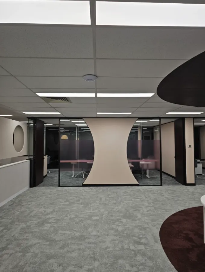 Modern office interior featuring ceiling solutions, beige walls and curved design elements, glass partitions with chairs and tables, and soft lighting creating a calm ambience.